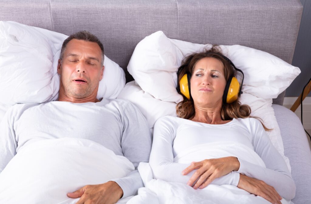 Couple lying in bed with the woman wearing hearing protection and glaring at her husband to her right because of his snoring.