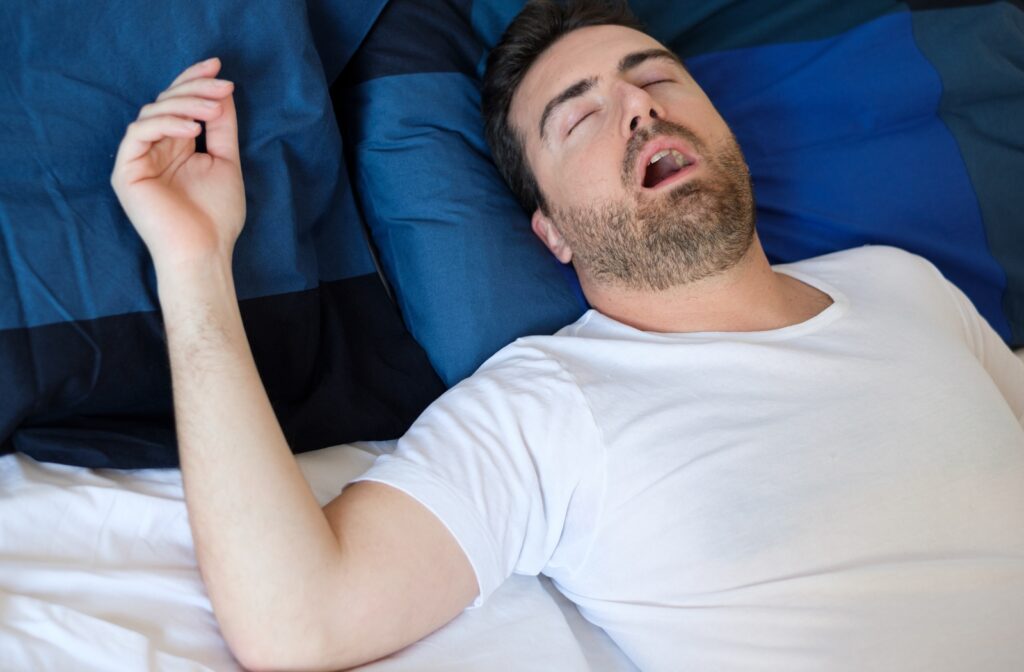 A man with a white shirt sleeping on his bed with his mouth wide open, snoring because he has untreated sleep apnea
