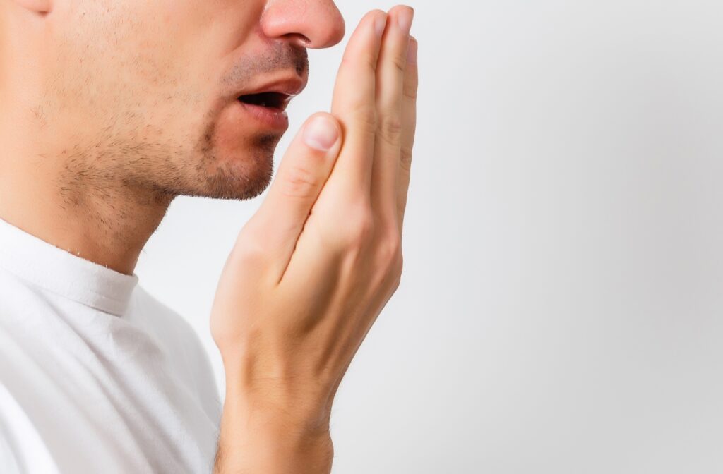 A man with a white shirt in front of a white background, holding his hand flat in front of his mouth to check if he has bad breath