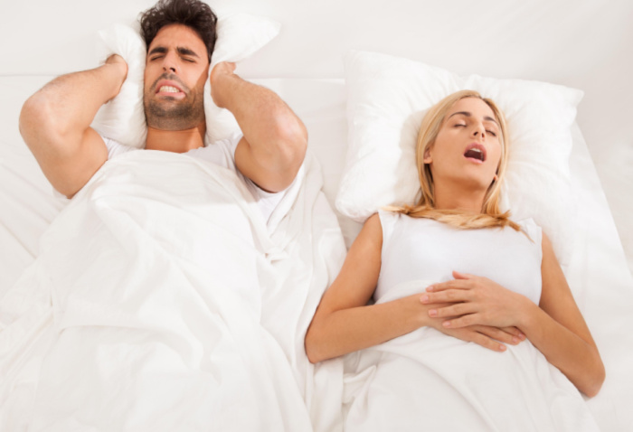 A woman sleeping beside her husband snoring extremely loudly keeping him awake as he tries to cover his ears with his pillow