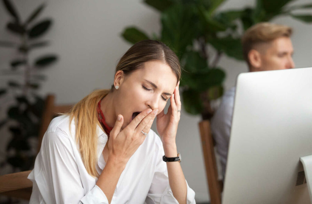 A woman sitting in front of her computer is yawning, feeling tired and sleepy during the day. A sign of possible  sleep apnea.