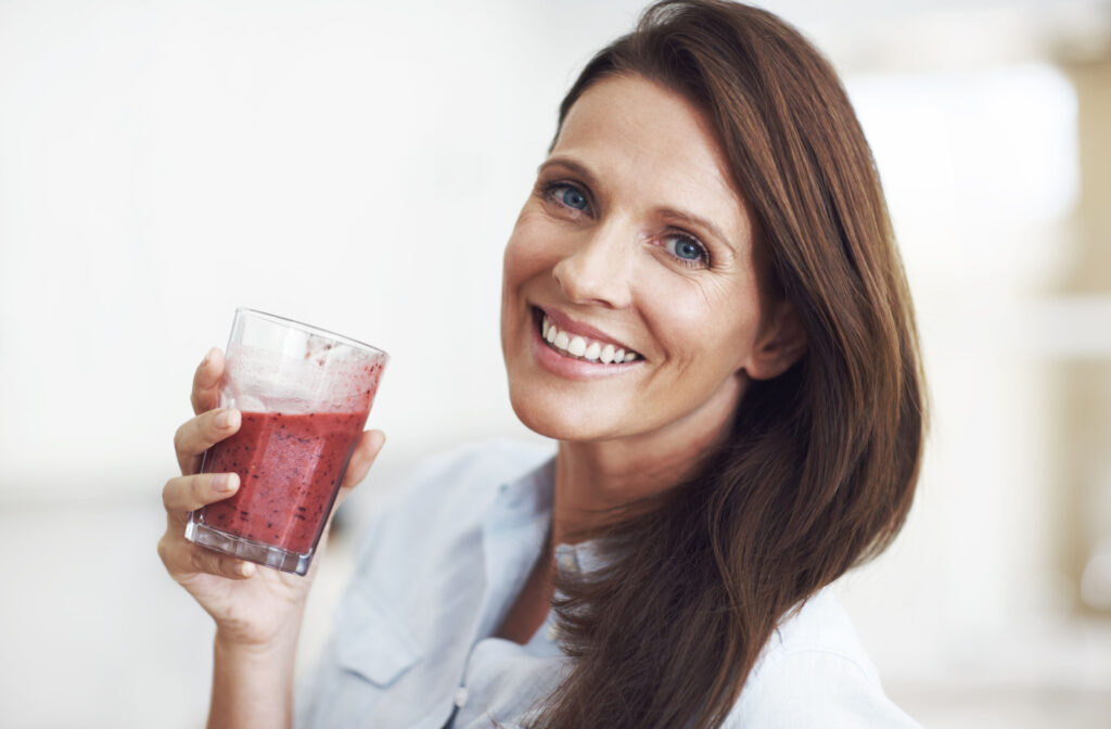 A woman holding a dairy-free fruit smoothie in her right hand as she smiles and looks directly at the camera.