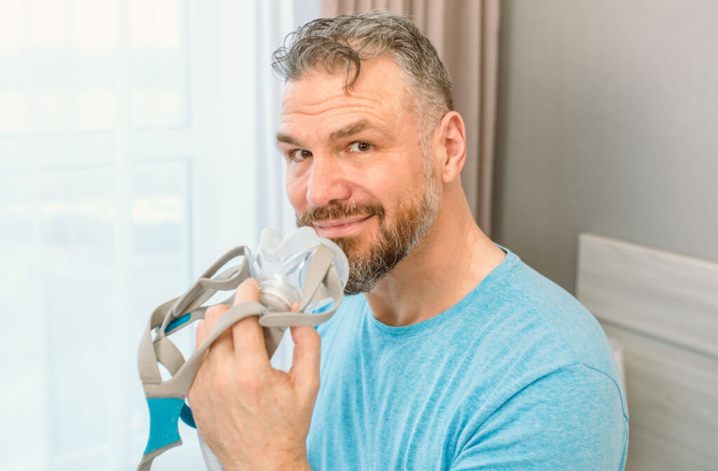 Close-up of a middle-aged man sitting up in bed and holding a CPAP machine up to his face, smiling.
