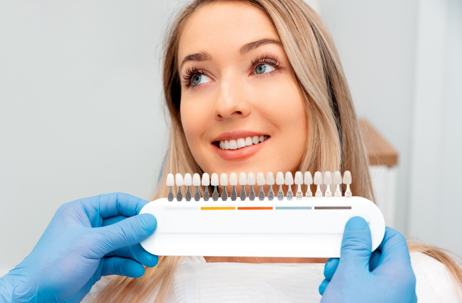A dentist holding up a veneer shade chart close to a female patient's teeth to help choose the right veneer shade for her.