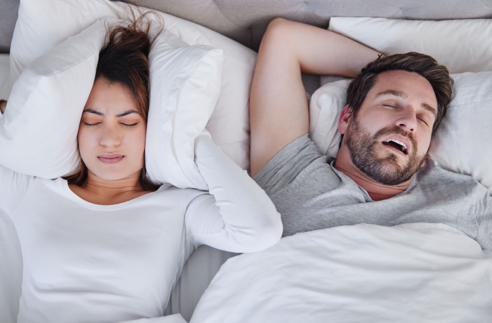 A person covers their ear with a pillow to not hear their partner snoring next to them.