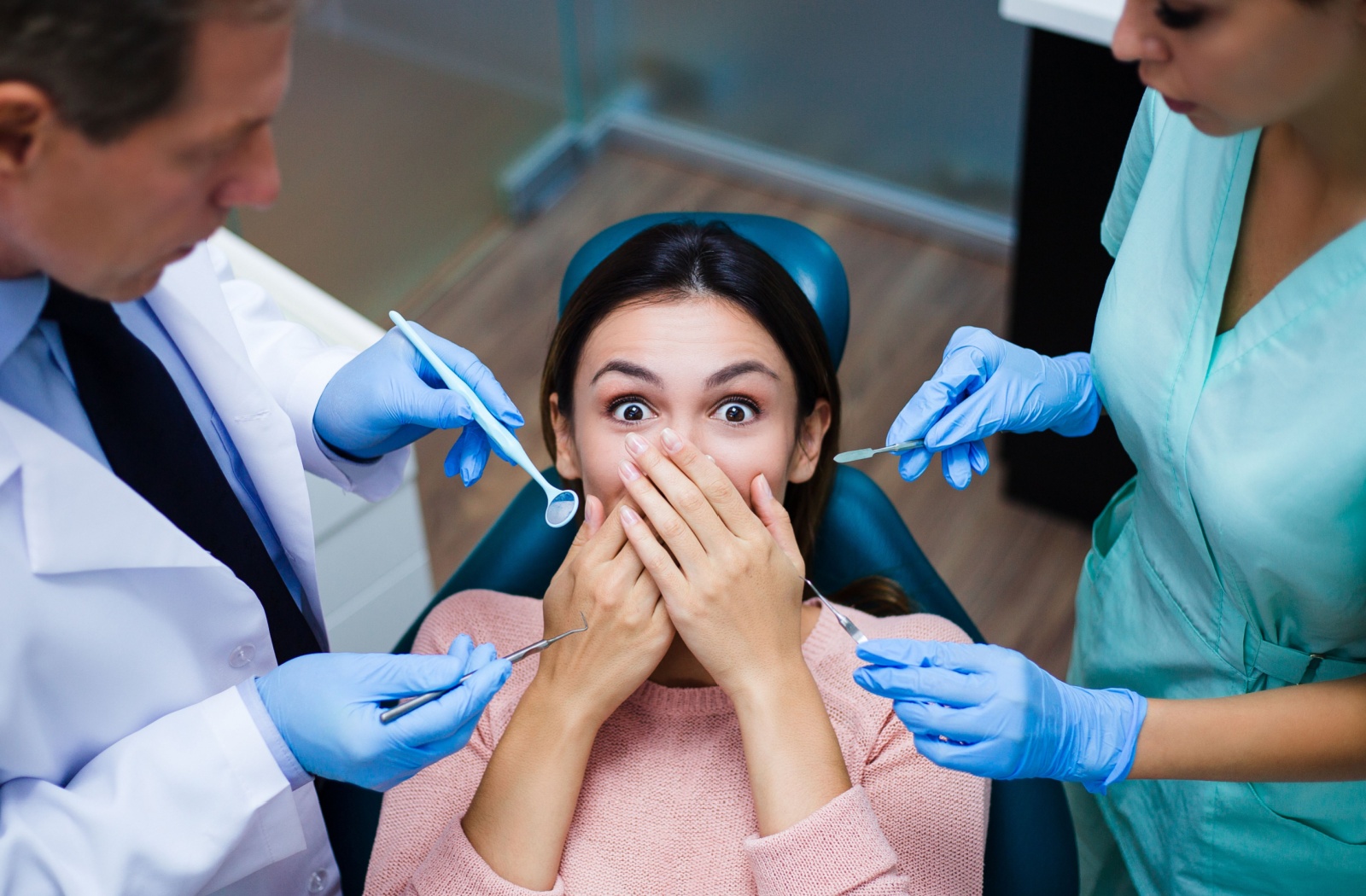 A patient with dental anxiety covers their mouth with their hands while a dentist and dental assistant stand over them holding dental tools.